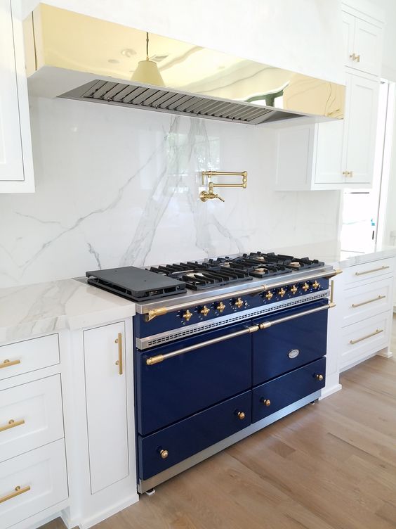 A white kitchen is spruced up with marble, a brass hood and a large cobalt blue cooker.