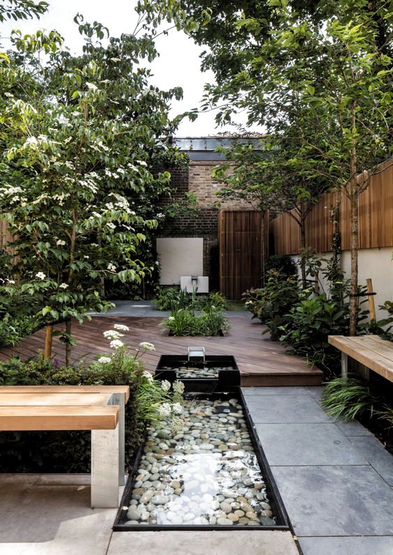 A beautiful summer terrace with trees, blooms and greenery, a fountain with pebbles, a bench and a deck.