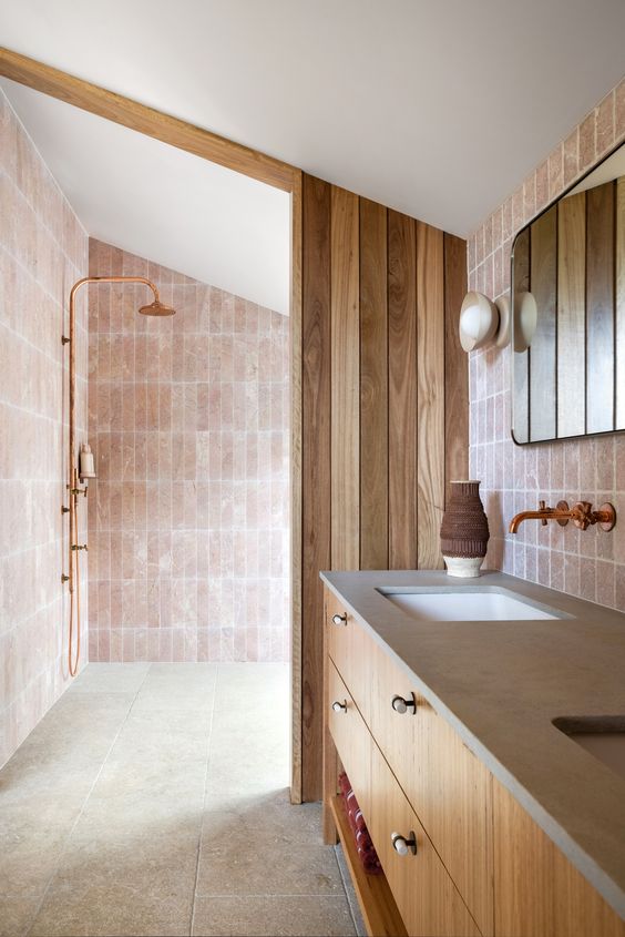 A blush bathroom clad with stone and skinny pink tiles, a double vanity, a wooden divider and some mirrors is cool.