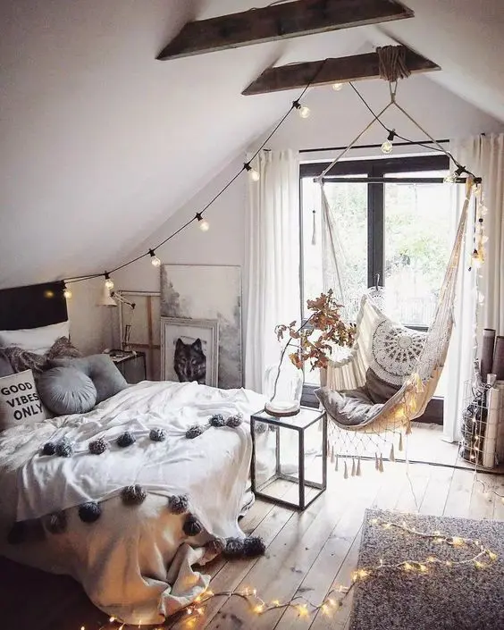 A boho teen attic bedroom with a bed with neutral bedding, a woven pendant chair, lights, wooden beams and artwork.
