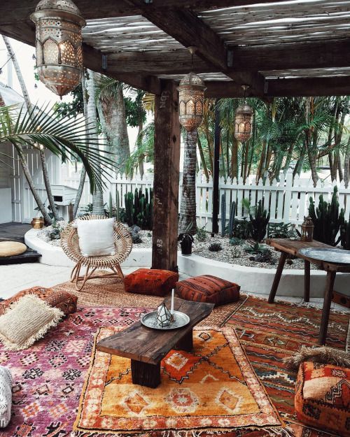 A boho terrace with colorful layered rugs, bright cushions and pillows, a rattan chair, Moroccan lanterns and cacti all around.
