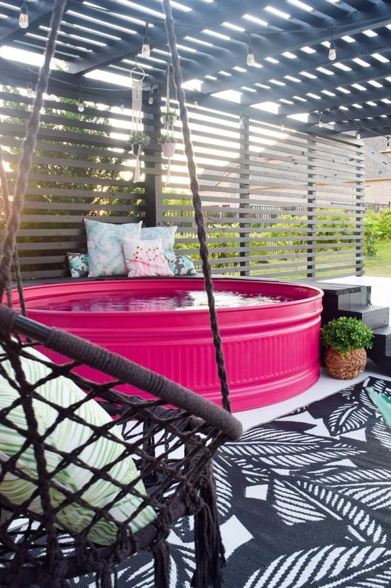 A bold patio with a hot pink stock tank pool, a black ladder, a woven chair with a pillow and some more pillows on the bench.