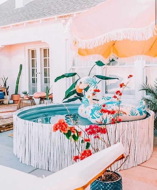 A bright and cool backyard with a wooden deck, a stock tank pool covered with whitewashed sticks, a colorful float, an umbrella and bold potted blooms.