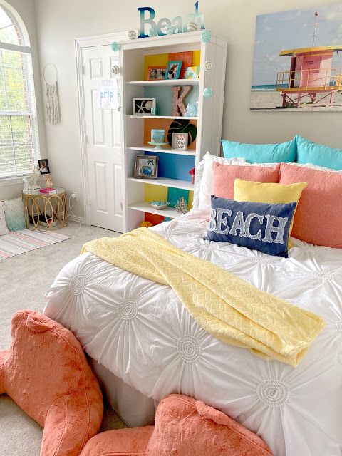 A bright beach teen girl room with a white storage unit, a bed with colorful bedding, coral pillows on the floor, a rattan table and a beach artwork.