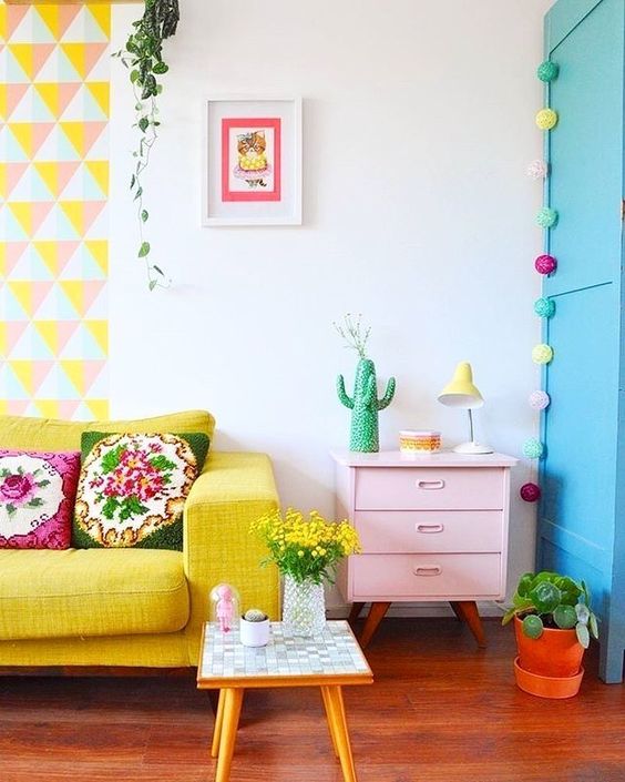 A bright geometric hanging, a yellow sofa, a pink dresser, colorful garlands and blooms and greenery in pots.