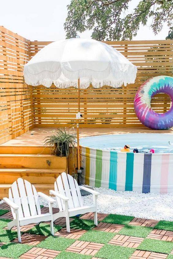A bright outdoor nook with a rainbow stock tank pool, a wooden deck, colorful floats, an umbrella, white chairs and a wooden rug.