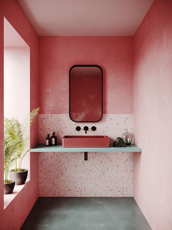A bright pink bathroom with a terrazzo accent, a terracotta sink on the vanity, potted greenery and a mirror in a black frame.