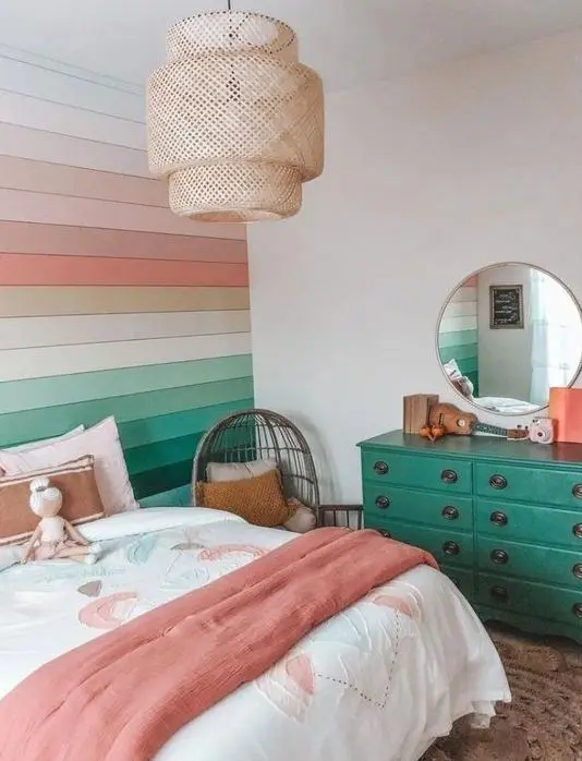 A bright rainbow teen girl room with a bold rainbow paneled wall, a bed with bright bedding, an emerald dresser, a round mirror and a woven pendant lamp.