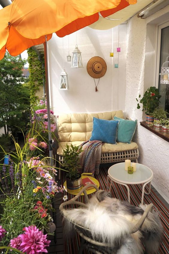 A bright summer balcony with bold textiles, rattan and metal furniture, colorful candle lanterns and an umbrella plus potted blooms.