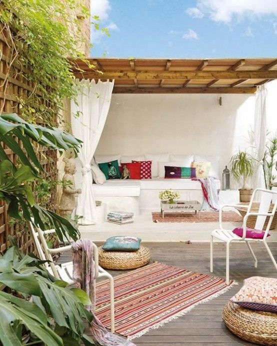 A bright summer terrace with a built-in bench, with bright pillows, rugs, wicker ottoman, potted greenery.