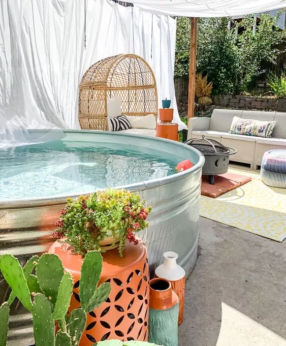 A catchy backyard with a stock tank pool, a sofa, a tent, an egg-shaped chair, a grill and some bright decor.