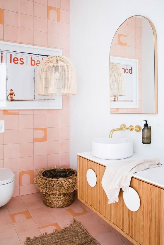 A catchy bathroom with pink tiles, a wicker lamp and basket, a floating vanity and gold fixtures for a glam touch.