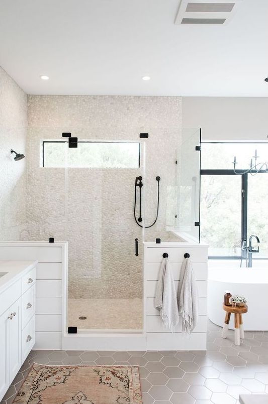 A chic neutral bathroom with grey hex tiles on the floor, faux pebbles in the shower space and planked pony walls and a large window.