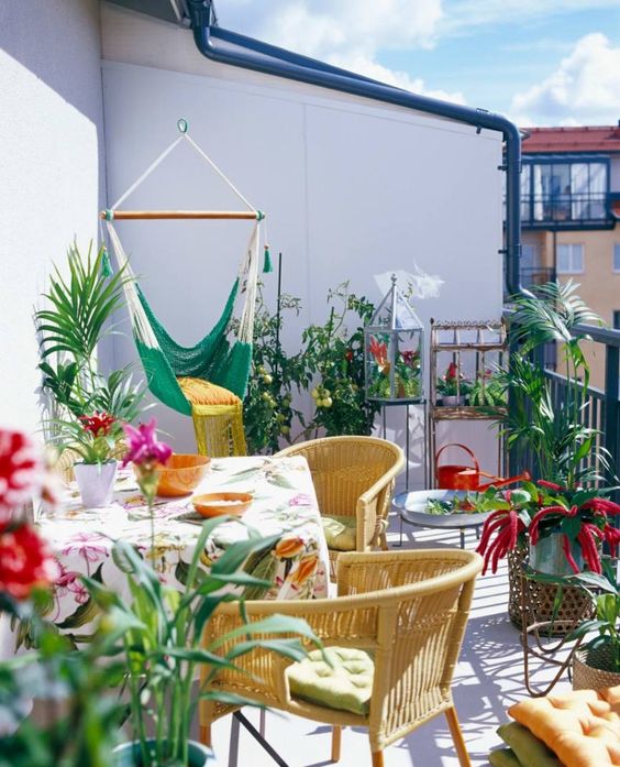 A colorful summer balcony with yellow chairs, printed textiles, a green hammock chair and lots of potted greenery and blooms.