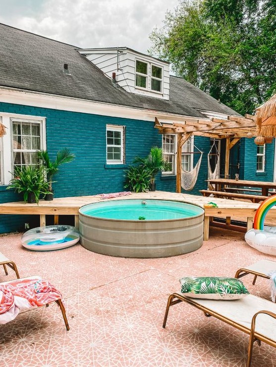 A cool and bright backyard with a wooden deck at the stock tank pool, loungers with colorful pillows, potted plants is welcoming.