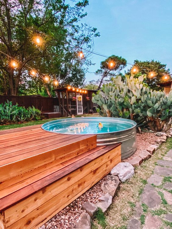 A cool outdoor space with a stock tank pool, some greenery and cacti around, a stepped wooden deck and string lights.