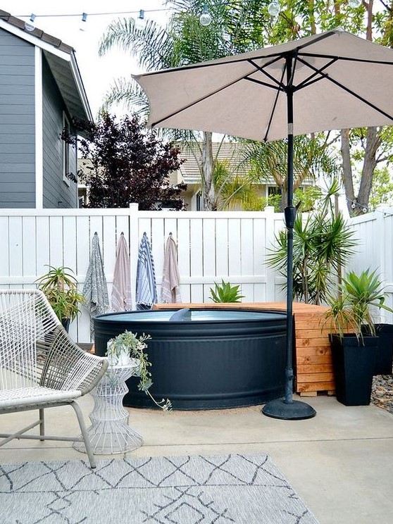 A cozy pool nook with a black stock tank pool, a wooden deck, potted plants, an umbrella and a woven chair and a side table.