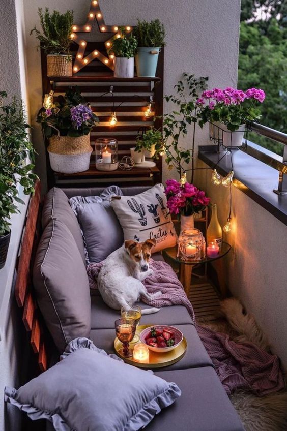 A cozy small balcony with a pallet wall and sofa, some potted blooms and greenery and candle lanterns.