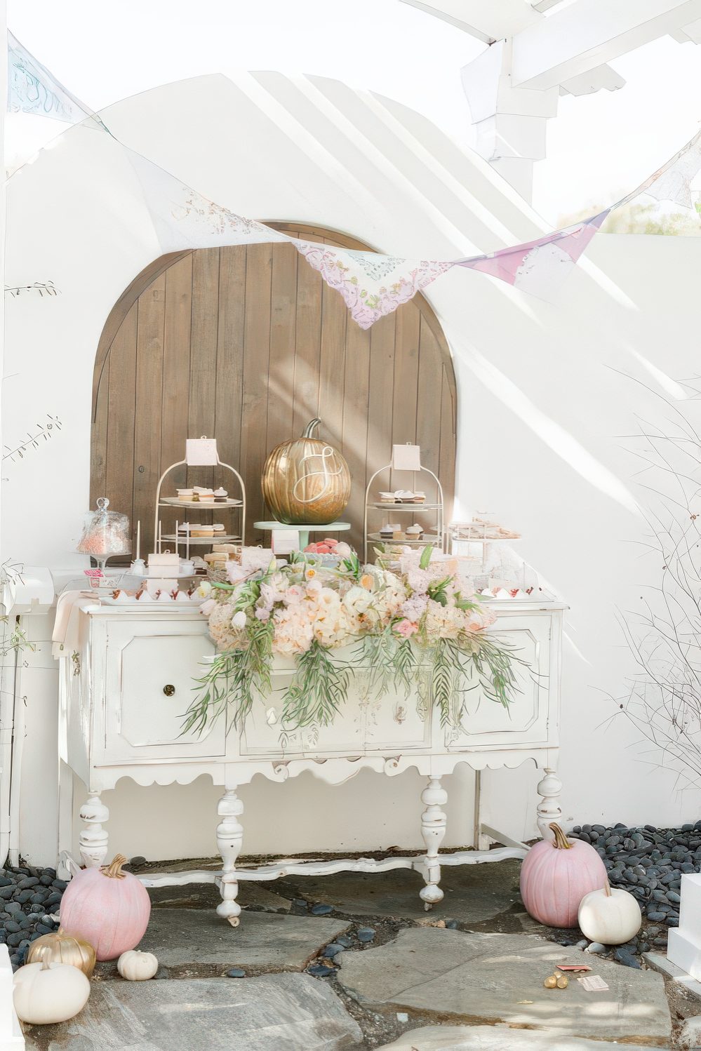 A delicate fall dessert table with a vintage sideboard, lots of painted pumpkins, a pink floral arrangement and a pink doily garland.