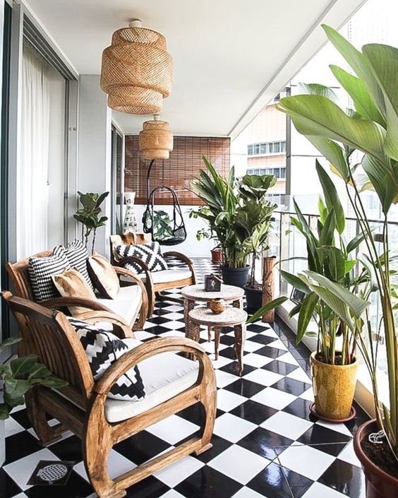 A large balcony with black and white tiles, wooden furniture, wicker lamps and printed pillows and lots of plants.