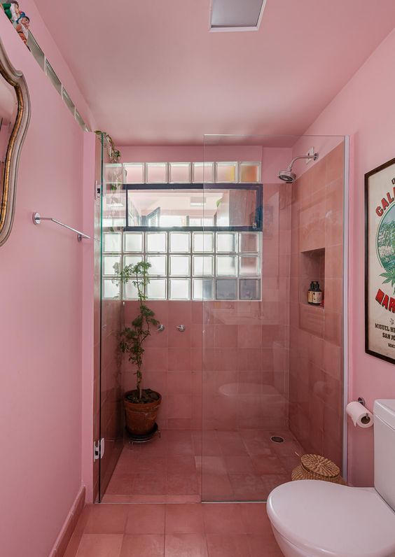 A light pink bathroom with coral tiles in the shower, a window and white appliances and greenery is cool and bright.