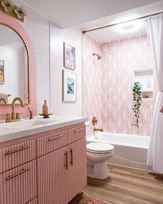A lovely pink bathroom with printed tiles, pink and white penny tiles, a pink fluted vanity, brass fixtures and potted greenery.
