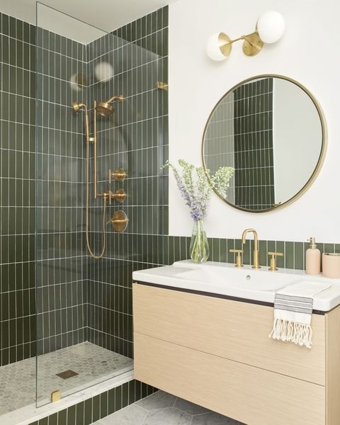 A modern bathroom with green skinny tiles, a floating vanity, a round mirror, gold and brass fixtures is cool.