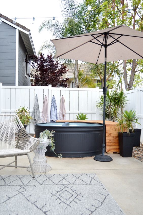 A modern stock tank pool in black, a wooden deck, an umbrella, potted greenery, a woven chair and a side table.