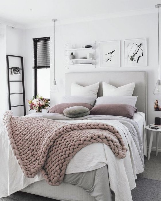 A neutral and pastel bedroom with a grey upholstered bed, layered blankets, black touches and bulbs hanging down.