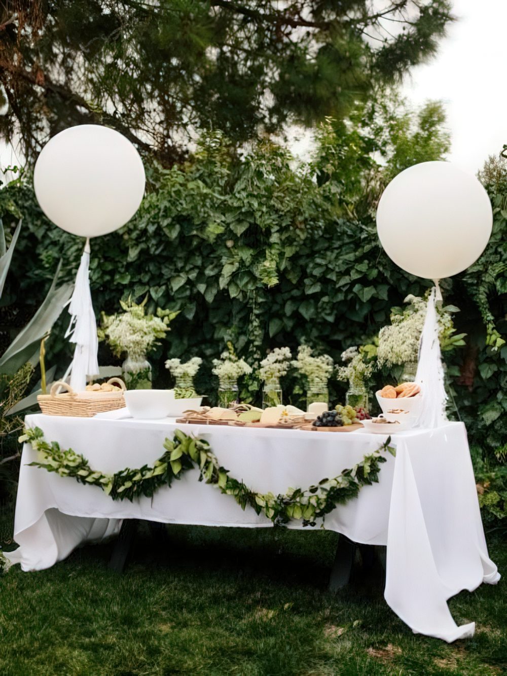 A neutral baby shower dessert table in white and green, with balloons and a greenery garland plus some neutral blooms.