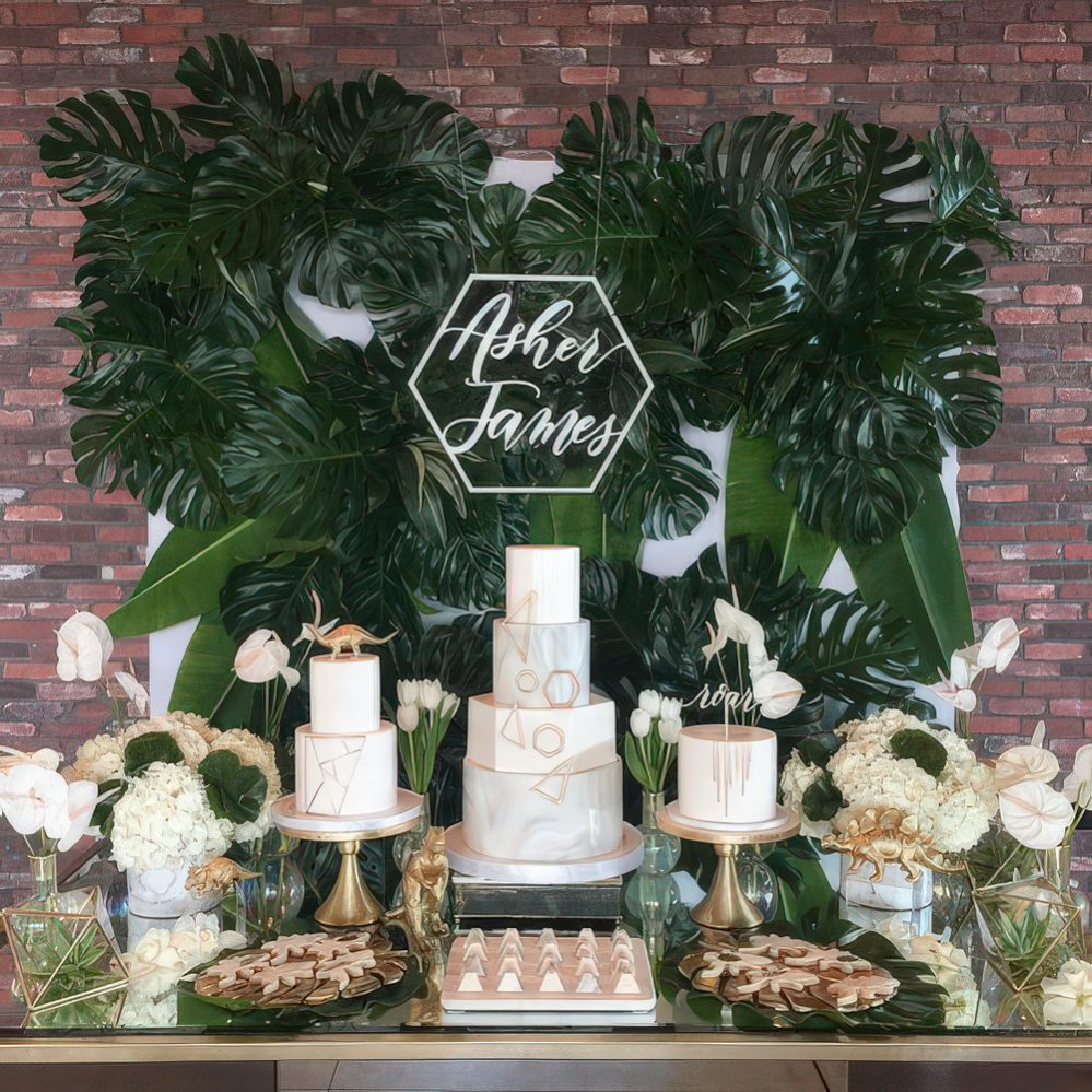 A neutral dessert table with a tropical feel, a lush tropical leaf wall, touches of gold, dinosaurs and white blooms.