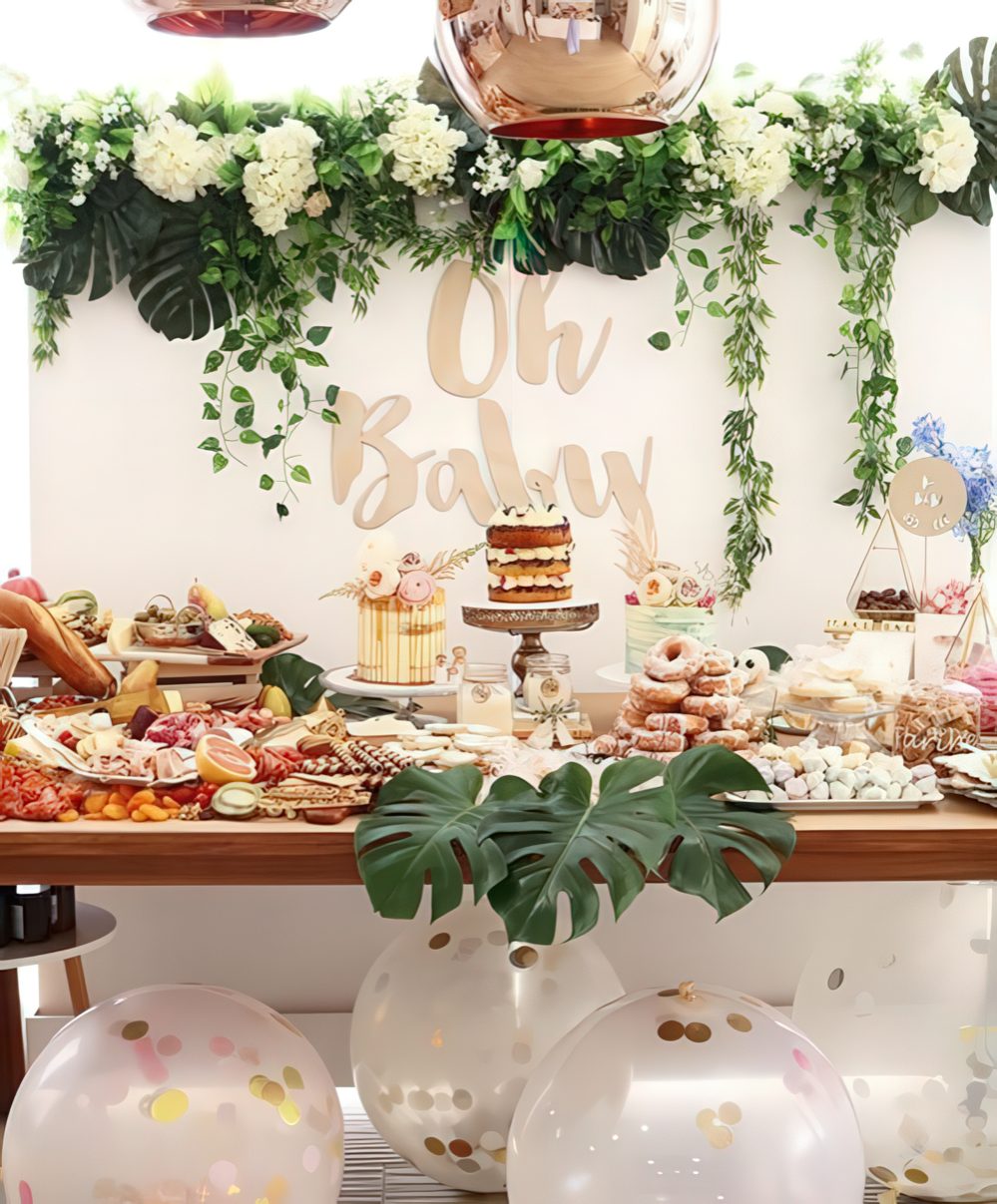A neutral dessert table with lush greenery and white bloom garlands, large balloons with confetti, tropical leaves.