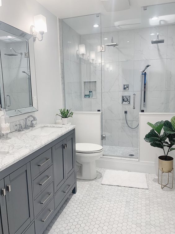 A neutral modern bathroom clad with marble hex tiles, with a shower space with half walls, a grey vnaity and a large mirror.