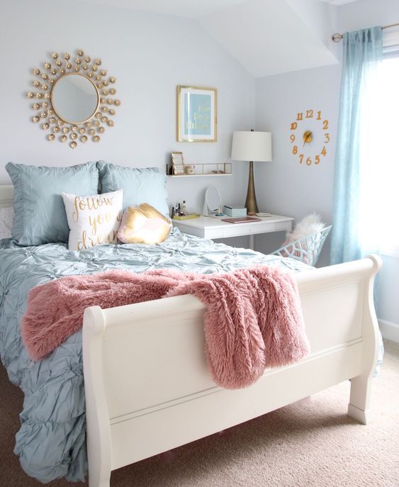 A pastel bedroom with a white bed and pastel bedding, blue curtains, a white desk as a nightstand, a white chair, a clock, a mirror and a table lamp.