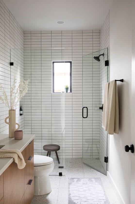 A peaceful and welcoming small bathroom clad with white skinny tiles, with a chic vanity, black fixtures and a small window in the shower.