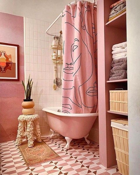 A pink and white bathroom with a pink free-standing tub, a printed curtain, geo tiles on the floor and some decor.