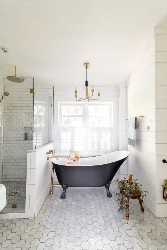 A refined farmhouse bathroom clad with hex and subway tiles, with a vintage bathtub, a stool with a plant and a vintage chandelier.