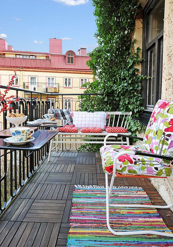 A simple and colorful balcony with bright textiles, some metal furniture and railing tables plus greenery.
