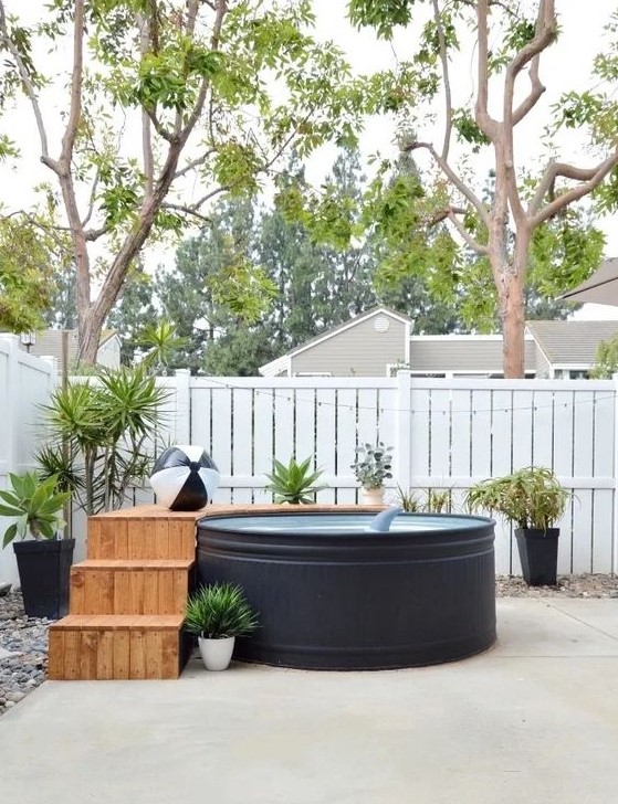 A simple and pretty backyard with a black stock tank pool, a wooden ladder and a deck, potted plants and fun floats is welcoming.
