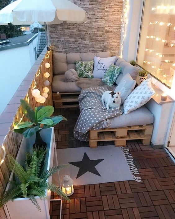 A small and cozy balcony with a pallet L-shaped sofa, printed pillows, plants in pots, lights and candle lanterns.