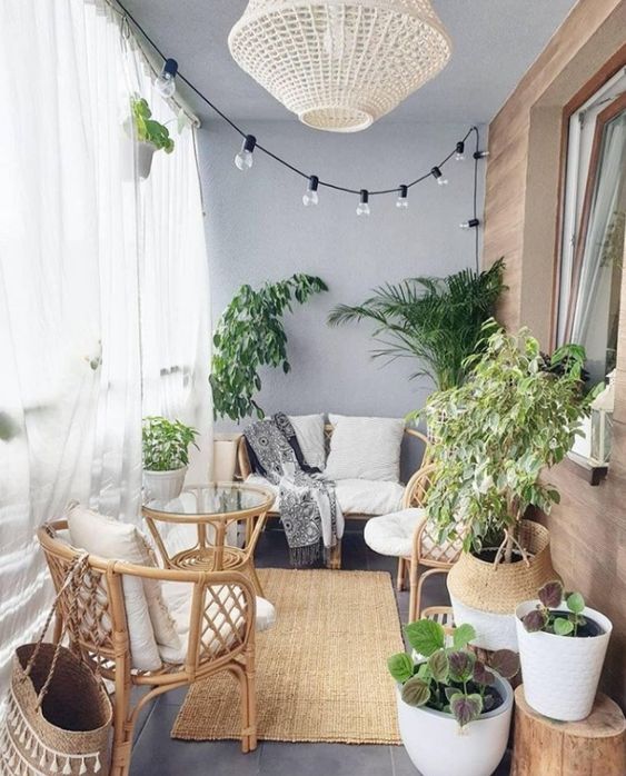 A small balcony with sheer curtains, lights, a wicker lamp, rattan furniture, statement plants and baskets is very cozy.
