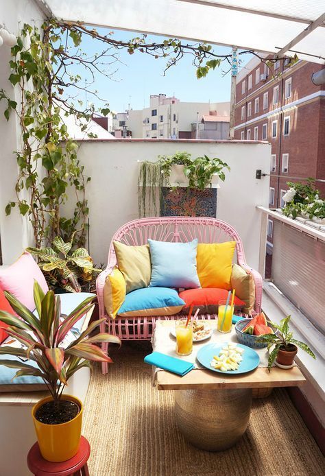 A small bright balcony with a pink loveseat, a small table, a white bench with storage, potted greenery and bright pillows.