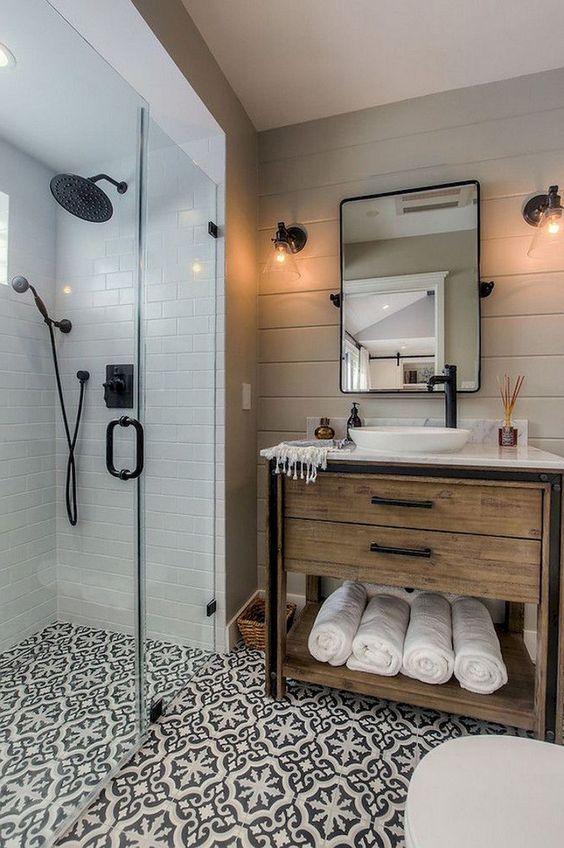 A small farmhouse bathroom with neutral tiles, patterned ones on the floor, a wooden vanity and black fixtures here and there.