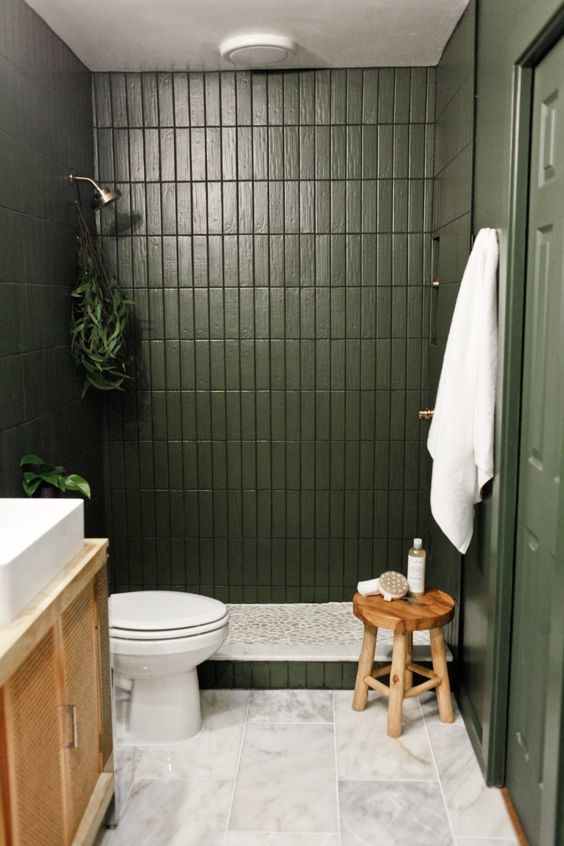 A small green bathroom with skinny tiles, a cane vanity, a wooden stool and some plants and white towels.