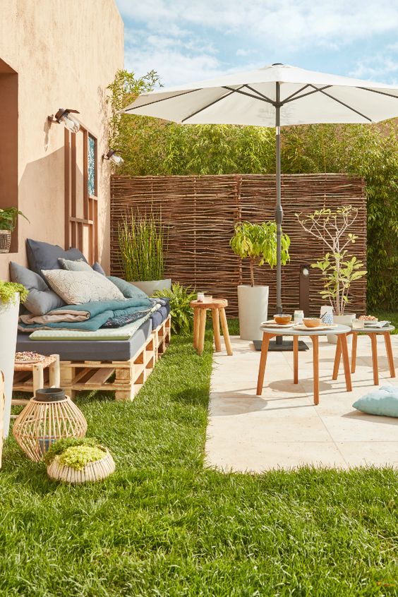 A small rustic summer terrace with a tiled space, a pallet sofa, wooden stools and potted greenery and candle lanterns.
