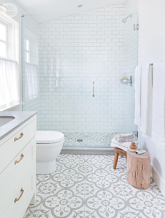 A small serene bathrooms with white subway tiles, patterned ones on the floor, wooden furniture and a window with a curtain.