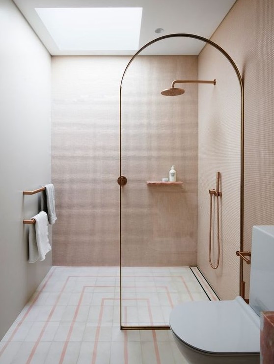 A small unique bathroom clad with blush mini tiles, with neutral and pink ones on the floor and a skylight over the shower plus brass touches.