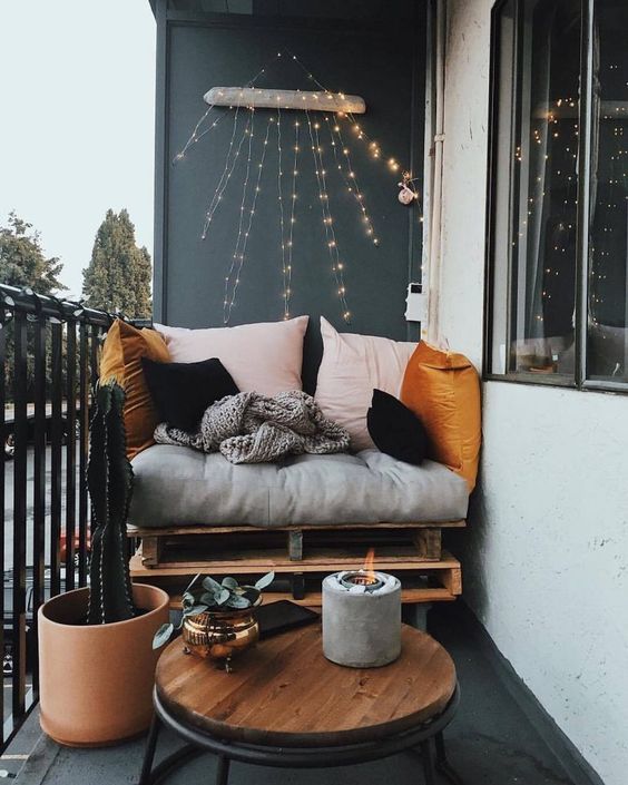 A small yet chic balcony with a pallet seat, colorful pillows, a table, a cactus and a candle in a concrete candleholder.