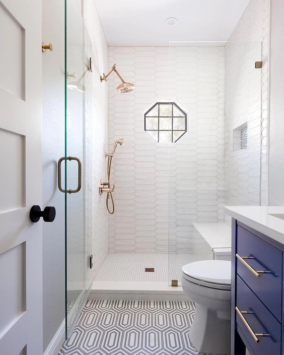 A small yet stylish bathroom with catchy long tiles in the shower and patterned ones ont he floor, with a bold blue vanity, brass touches and a geometric window.