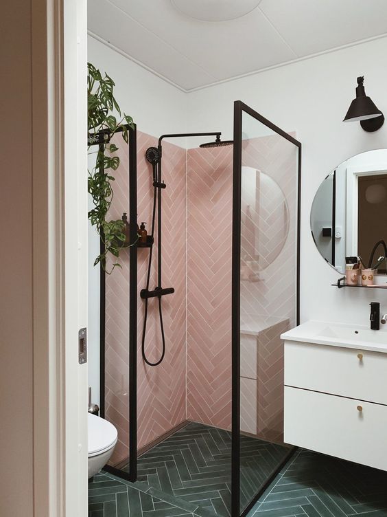 A stylish small bathroom with green herringbone tiles on the floor, pink ones in the shower, a floating vanity and black framing.
